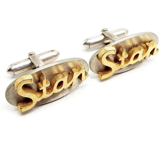 Front view of the Swank Mid Century vintage name cufflinks. They are silver tone color ovals with a script style font cut out name Stan on the front in gold tone color metal.