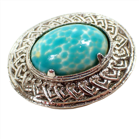 Angled front view of the retro vintage Jacobite Celtic brooch pin. The metal is silver tone in color. It is oval in shape with a Celtic V design all the way around the edge. There is a prong set fancy glass cab in the middle that is off white yellow with speckled spots of aqua blue color.