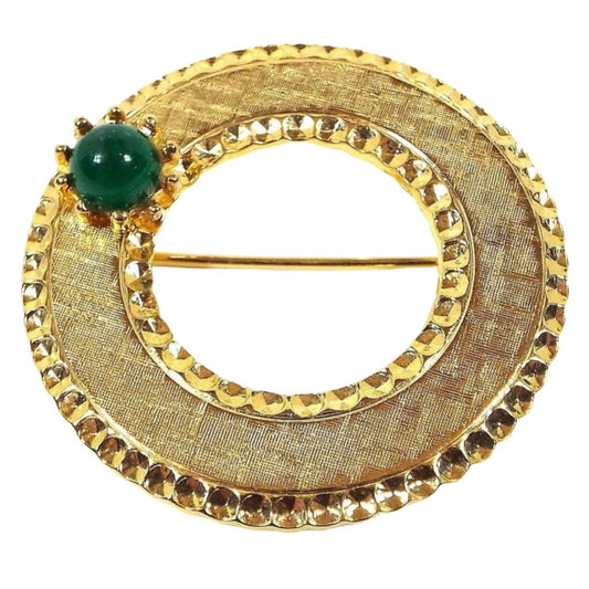 Front view of the retro vintage Beaujewels brooch. It is gold tone in color. It has an open middle area. There metal is textured brushed and there is a stamped faceted style outer and inner edge. There is a small green lucite cab on one side.