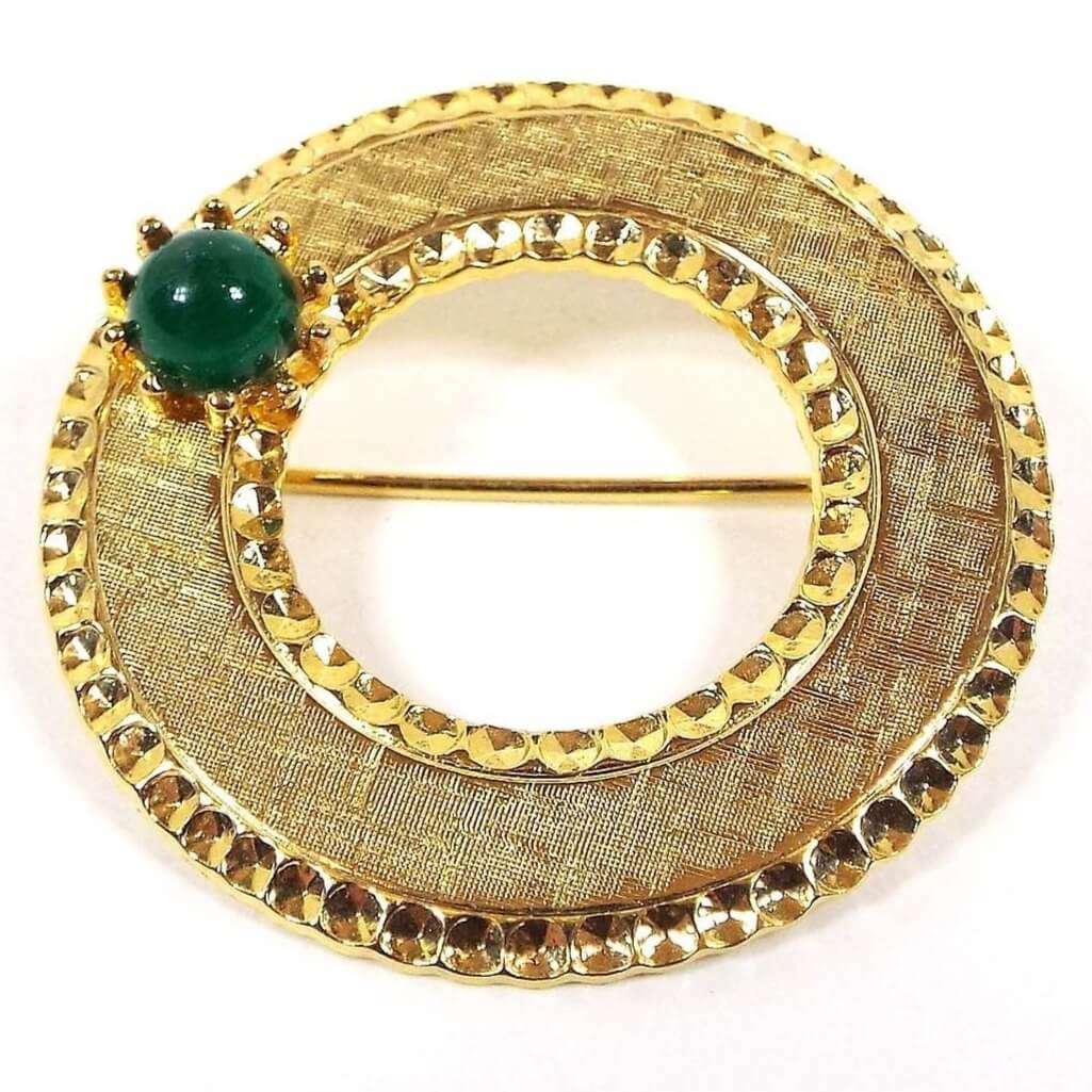 Front view of the retro vintage Beaujewels brooch. It is gold tone in color. It has an open middle area. There metal is textured brushed and there is a stamped faceted style outer and inner edge. There is a small green lucite cab on one side.