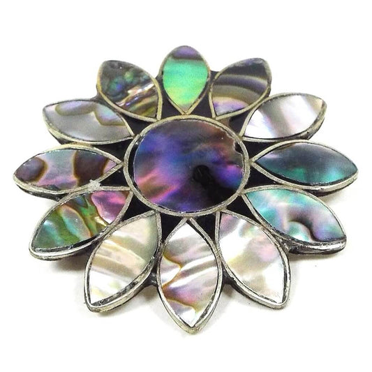 Front view of the retro vintage Mexican Alpaca flower brooch pin. The metal is silver tone in color. it is shaped like a sunflower with a circle in the middle and marquis shaped petals all the way around. Each part of the flower has an inlaid piece of abalone shell that is multi colored with flashes of color as you move around in the light.