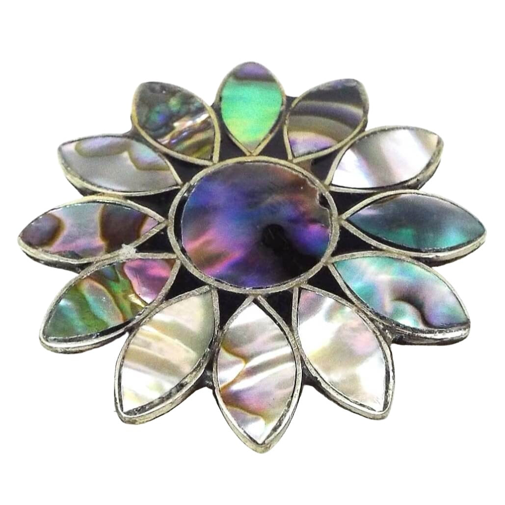 Front view of the retro vintage Mexican Alpaca flower brooch pin. The metal is silver tone in color. it is shaped like a sunflower with a circle in the middle and marquis shaped petals all the way around. Each part of the flower has an inlaid piece of abalone shell that is multi colored with flashes of color as you move around in the light.