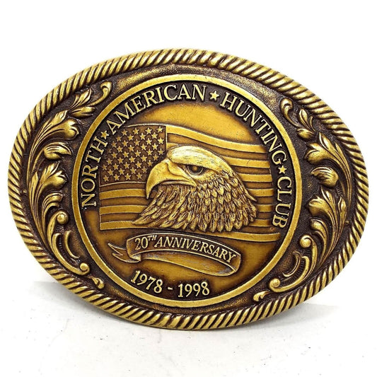 Front view of the retro vintage North American Hunting Club belt buckle. It is oval and antiqued brass in color. The sides have a raised leaf design. The middle has an oval with the wording for the 20th Anniversary 1978-1998 and has an eagle's head in front of the American Flag.