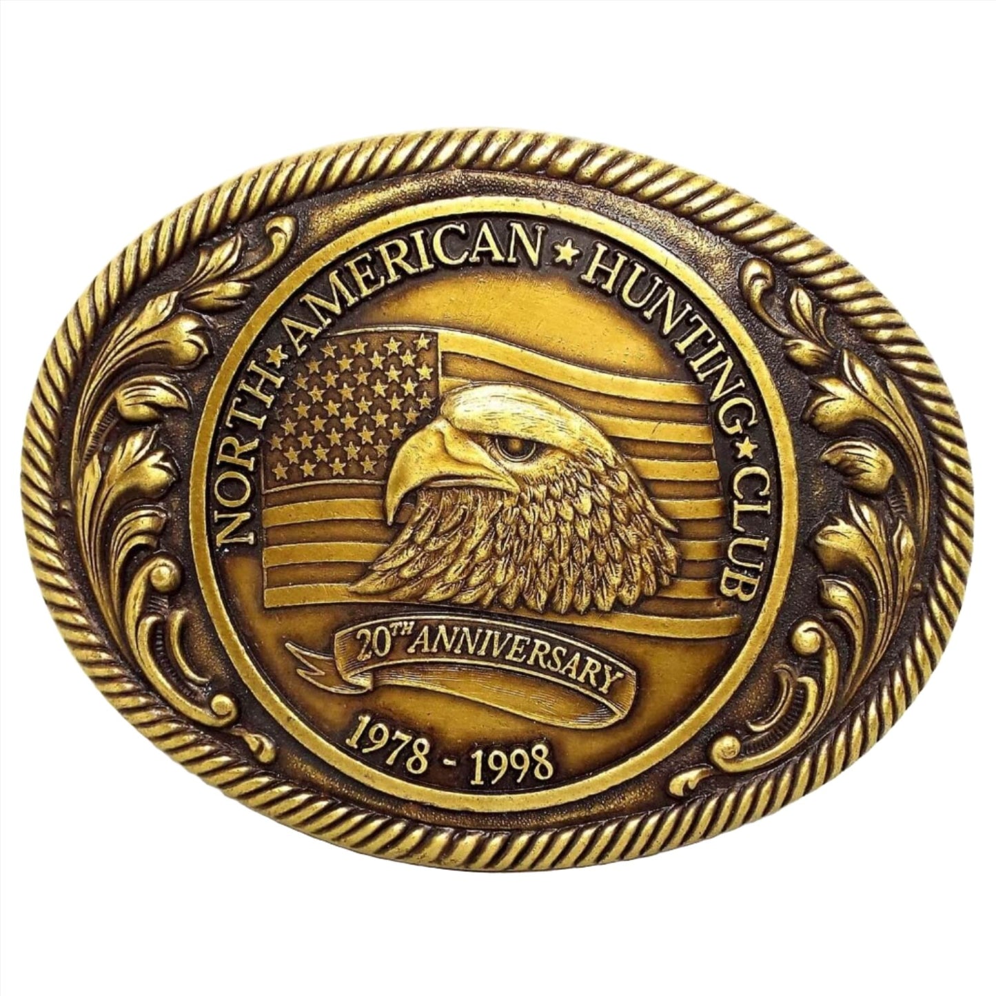 Front view of the retro vintage North American Hunting Club belt buckle. It is oval and antiqued brass in color. The sides have a raised leaf design. The middle has an oval with the wording for the 20th Anniversary 1978-1998 and has an eagle's head in front of the American Flag.