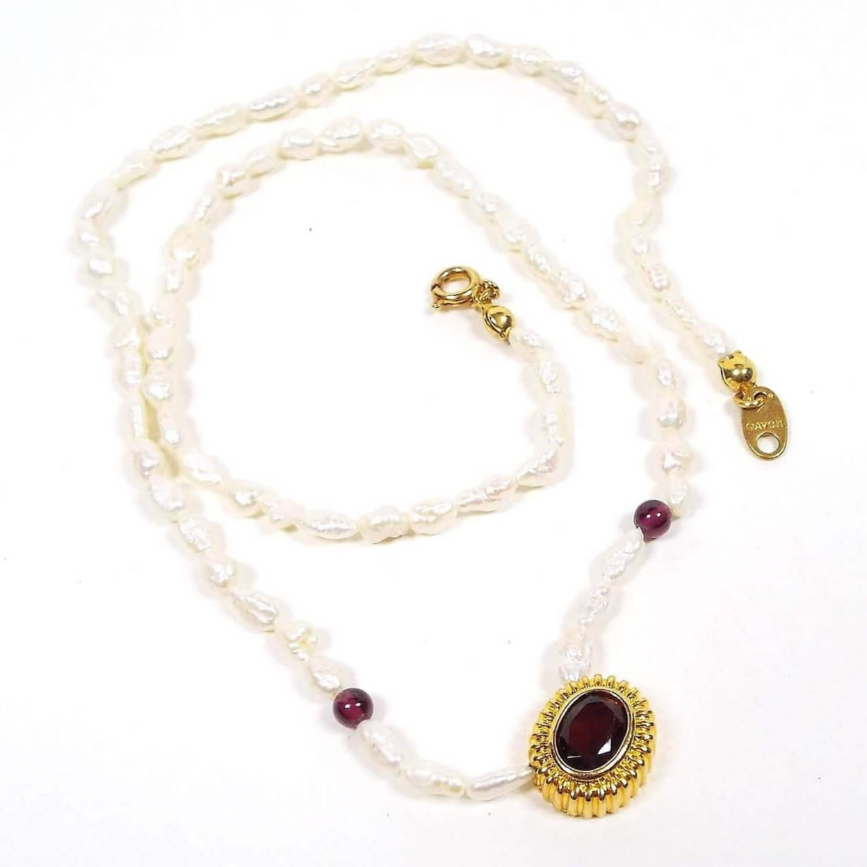 Top view of the retro vintage Avon garnet and freshwater cultured pearl beaded pendant necklace. The metal around the pendant and clasp is gold tone in color. The necklace is beaded with small rice pearls and two small round garnet beads. The bottom pendant has an oval garnet cab in the middle. 