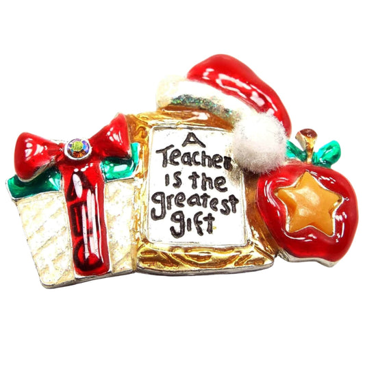 Front view of the retro vintage AMJC teacher brooch pin. It is enameled in red, green, and gold color. There is a present with a bow that has ABC on it, an apple with a star on it, a frame that says a Teacher is the greatest gift, and a Santa hat on top with glitter and a fabric pom pom.