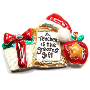 Front view of the retro vintage AMJC teacher brooch pin. It is enameled in red, green, and gold color. There is a present with a bow that has ABC on it, an apple with a star on it, a frame that says a Teacher is the greatest gift, and a Santa hat on top with glitter and a fabric pom pom.
