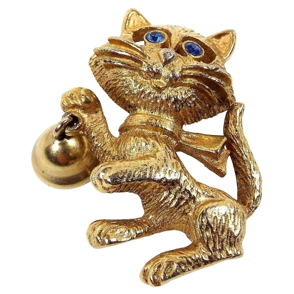 Front view of the Avon retro vintage cat and bell brooch. It is gold tone in color and is textured. The cat is standing on its hind legs and is holding a bell. There are two blue rhinestones for the eyes.
