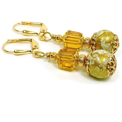 Side view of the handmade glass drop earrings. The metal is gold plated in color. There is a faceted orange cube bead at the top. The bottom round glass bead is off white with splashes of metallic gold color.