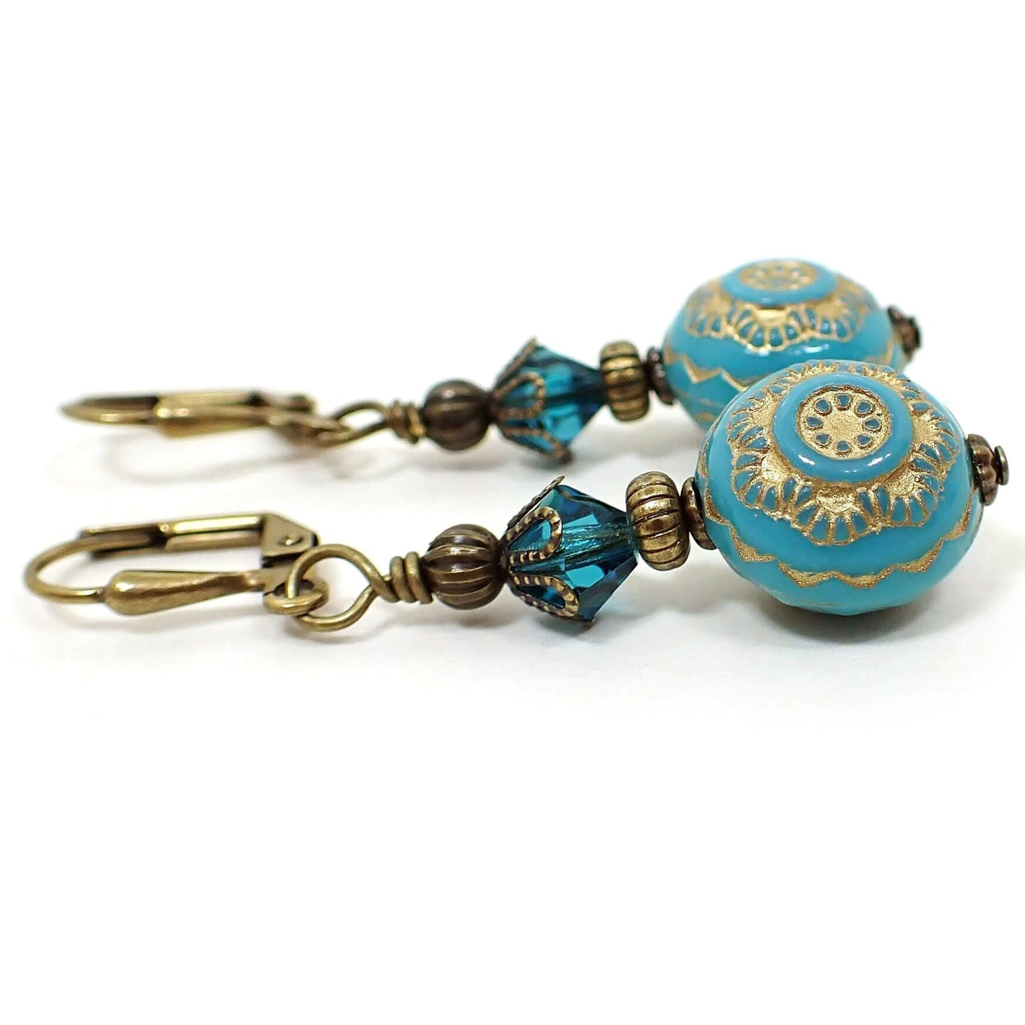 Side view of the handmade floral drop earrings. The metal is antiqued brass in color. There are faceted glass crystal beads at the top in a teal blue color. The bottom beads are turquoise blue in color and have a metallic antiqued gold painted flower design on them.
