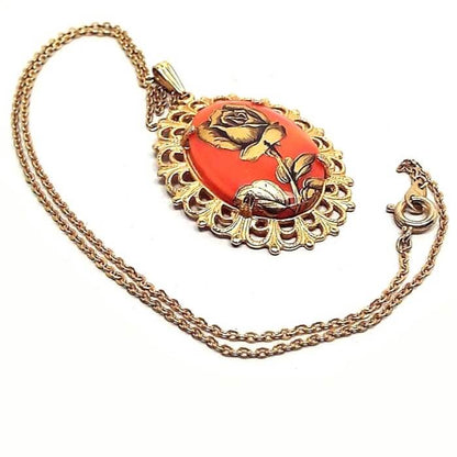 Front view of the Mid Century vintage floral pendant. The metal is gold tone in color. There is a cable chain with a spring ring clasp and oval pendant at the bottom. The pendant has a filigree setting with a bright orange glass cab. There is a black and antiqued gold color decal of a rose flower on top of the glass cab.