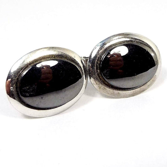 Front view of the Mid Century vintage faux hematite cufflinks. They are oval in shape and have silver tone color metal. The fronts have dark metallic gray glass cabs. You can see my hands and a light reflecting off them in the photo.
