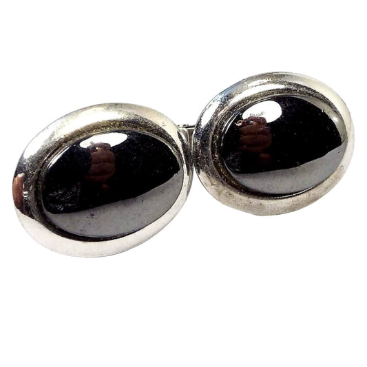 Front view of the Mid Century vintage faux hematite cufflinks. They are oval in shape and have silver tone color metal. The fronts have dark metallic gray glass cabs. You can see my hands and a light reflecting off them in the photo.