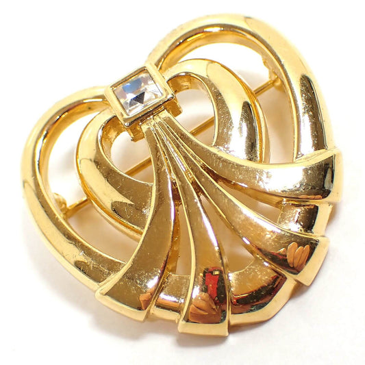 Angled front view of the retro vintage rhinestone heart brooch pin. The metal is shiny gold tone in color. It has an open heart style shape with four bands flaring out to the bottom of the heart. At the top is a single square clear rhinestone. You can see reflections of the camera and my hands on the bottom of the brooch.