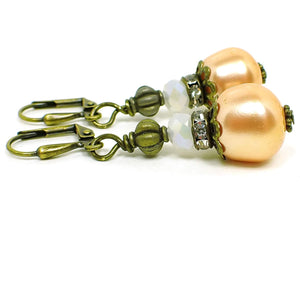 Side view of the handmade earrings made with vintage faux pearl beads. The metal is antiqued brass in color. There are faceted glass white crystal beads at the top, a spacer bead with clear rhinestones all the way around it, and the bottom beads are plastic faux pearl in a peach color. The peach beads are a larger potato style shape.