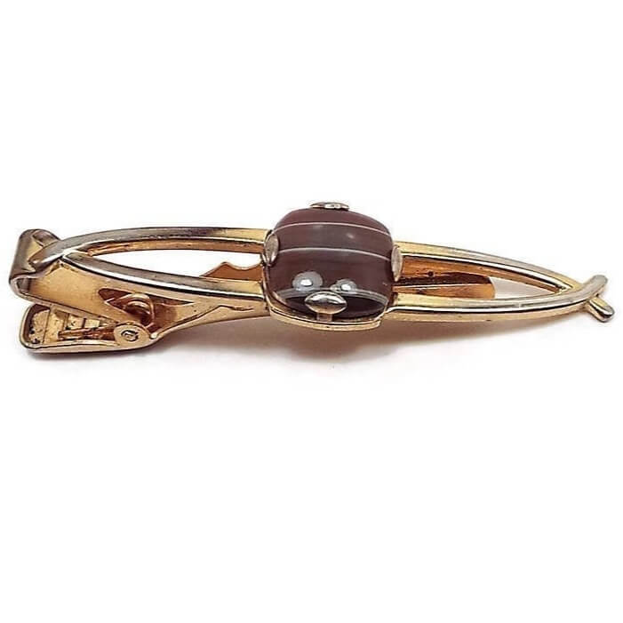 Front view of the Anson Mid Century vintage tie clip. The metal is gold tone in color. It has a curved line of metal on each side leaving the middle area open. In the very middle is a prong set fancy glass cab that is rounded square in shape and has shades of brown with thin white stripes going through it.