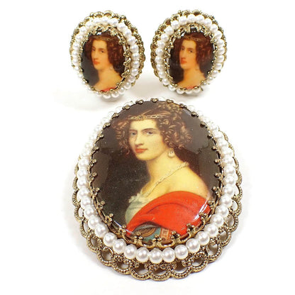 Angled front view of the Mid Century vintage  cameo set from West Germany. At the top of the photo is the pair of clip on earrings. There is a large brooch pin beneath them. All pieces have antiqued gold tone metal, oval shape with a filigree edge, and plastic faux pearl trim. There are plastic oval cameos with decals over them of a woman with curly hair and a red and white dress. 