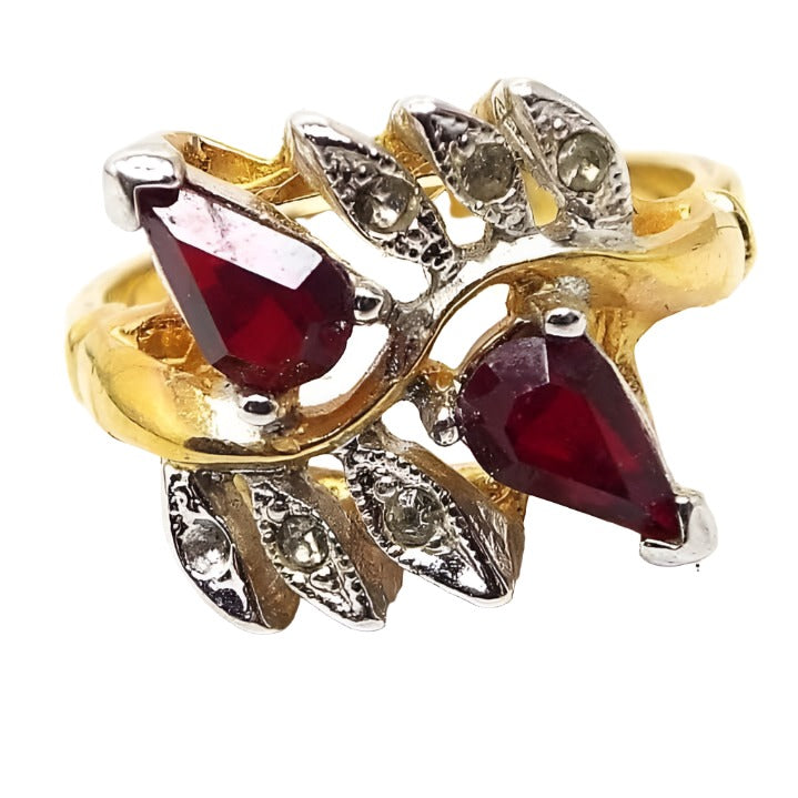 Front view of the retro vintage Vargas rhinestone cocktail ring. The metal is gold tone in color n the band and most of the setting. There are two teardrop shaped prong set red rhinestones angled away from each other on the top of the ring. On each side of those are three leaf shaped settings for small round adhesive set clear rhinestones with silver tone color on the front of the settings. 