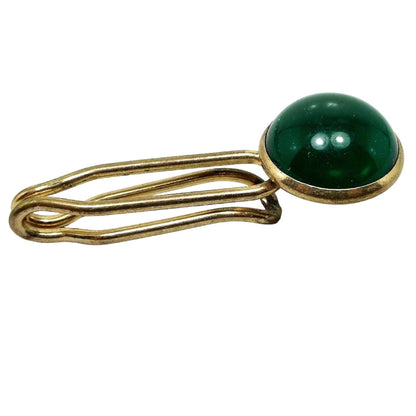 Angled front and side view of the Mid Century vintage lucite tie bar. The metal is gold tone in color. The bar is thick wire style with open slide on design. There is a round green lucite cab at the end.
