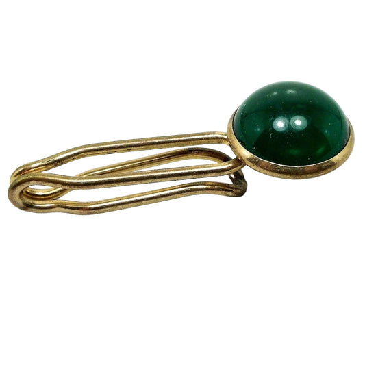 Angled front and side view of the Mid Century vintage lucite tie bar. The metal is gold tone in color. The bar is thick wire style with open slide on design. There is a round green lucite cab at the end.