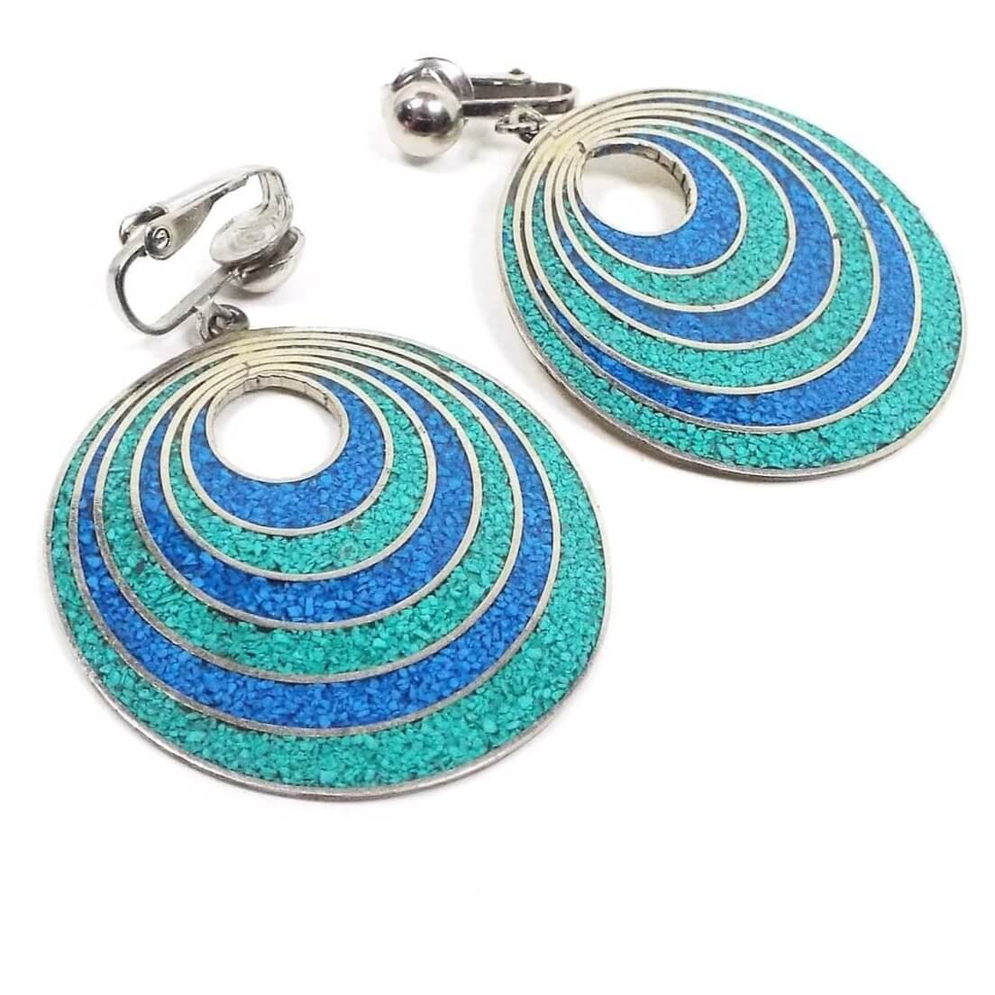 Front view of the retro vintage Mexican alpaca earrings. The metal is silver tone in color. There are clips at the top and large round drops at the bottom with a small round opening. There are rings of blue and green resin with tiny stone chips in them.