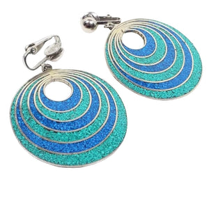 Front view of the retro vintage Mexican alpaca earrings. The metal is silver tone in color. There are clips at the top and large round drops at the bottom with a small round opening. There are rings of blue and green resin with tiny stone chips in them.