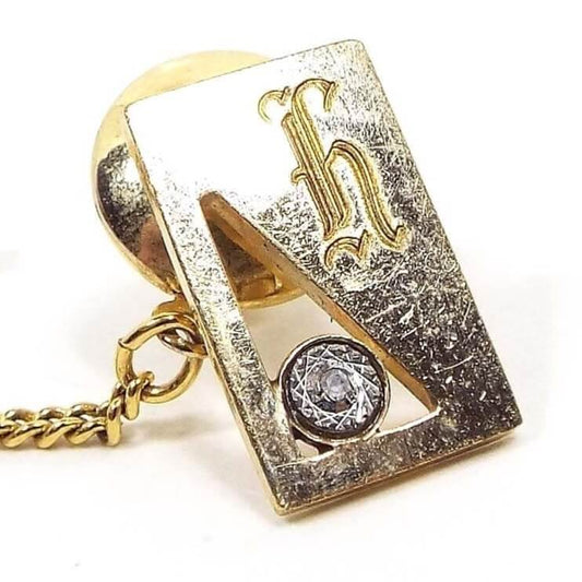 Front view of the Mid Century vintage gold filled initial tie tack. It is rectangle in shape and gold tone in color. There is a triangle cut out area on the left with a small round etched area that has a diamond chip in it. On the top right is a fancy letter H. The gold plated clutch back has a chain on it.
