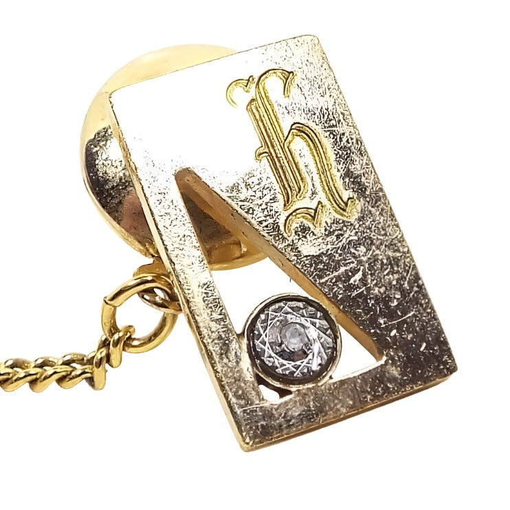 Front view of the Mid Century vintage gold filled initial tie tack. It is rectangle in shape and gold tone in color. There is a triangle cut out area on the left with a small round etched area that has a diamond chip in it. On the top right is a fancy letter H. The gold plated clutch back has a chain on it.