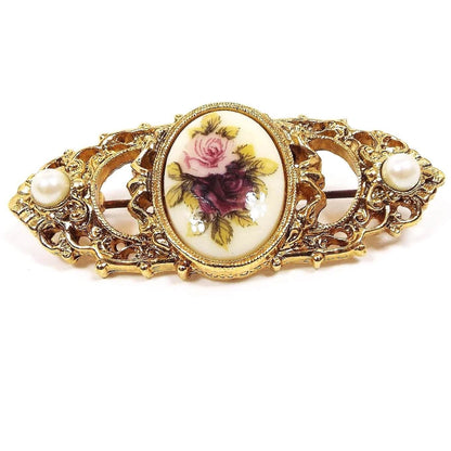 Front view of the retro vintage filigree floral brooch pin. The metal is gold tone in color. It has a filigree design with points at the ends and an oval in the middle. The ends have a plastic faux pearl on each side. The oval in the middle has a white plastic cab with a rose flower design painted on it. One flower is pink and the other is purple and they are surrounded with green leaves.