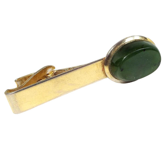 Front view of the retro vintage jade tie clip. The metal is gold tone in color. There is some rub wear at the end where some of the lighter base color is showing through from age. The other end has a thick oval jade gemstone cab that is dark green with a few darker color spots.