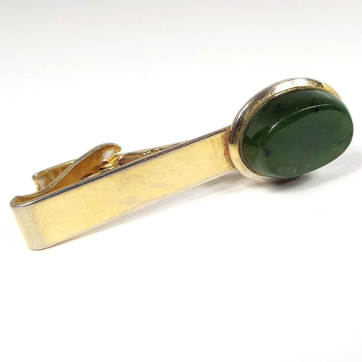Front view of the retro vintage jade tie clip. The metal is gold tone in color. There is some rub wear at the end where some of the lighter base color is showing through from age. The other end has a thick oval jade gemstone cab that is dark green with a few darker color spots.