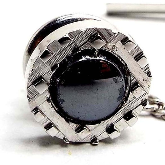 Close up view of the retro vintage faux hematite tie tack. It is round and the metal is silver tone in color. There is a metallic dark gray cab in the middle surrounded by a cut tiny square pattern around the edge.