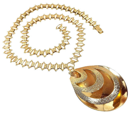 Top view of the Mid Century Vintage Sarah Coventry large teardrop pendant necklace. The chain is gold tone with fancy open links and textured pattern links. There is a snap lock clasp on the end. The pendant is a large teardrop with a cut areas on the front and a spring ring clasp at the top so you can remove it from the chain if you want.