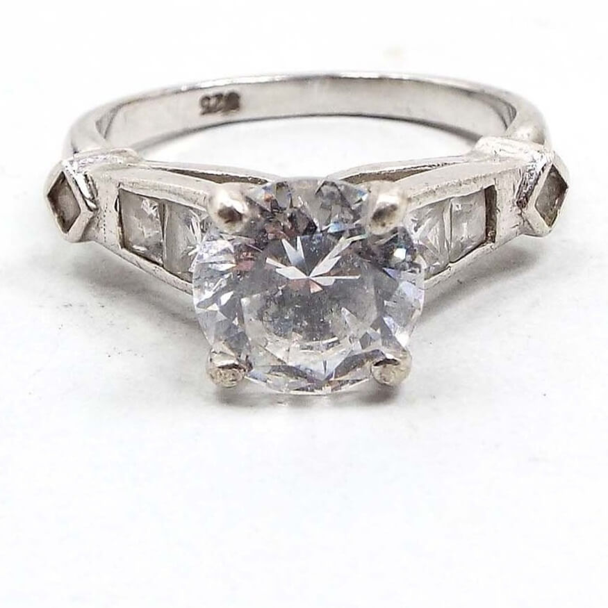 Front view of the retro vintage sterling silver cubic zirconia geometric ring. The top of the ring has a prong set round CZ stone in the middle. There are two channel set square stones on each side of it and a bezel set square stone set diagonally for a diamond shape on each side. 925 is marked on the inside of the ring.