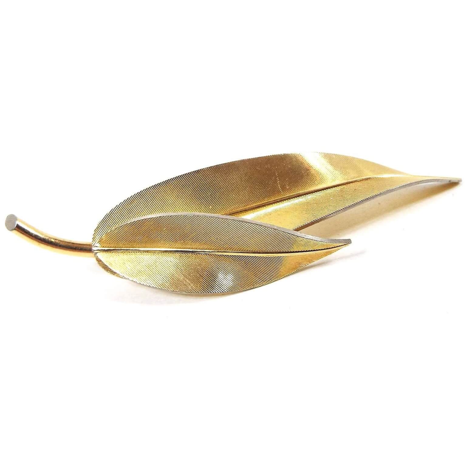 Front view of the Mid Century vintage Krementz leaf brooch pin. It is gold tone in color with a textured fine line front. There is a leaf stem holding two elongated leaves that curve at the end.