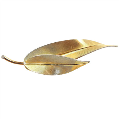 Front view of the Mid Century vintage Krementz leaf brooch pin. It is gold tone in color with a textured fine line front. There is a leaf stem holding two elongated leaves that curve at the end.