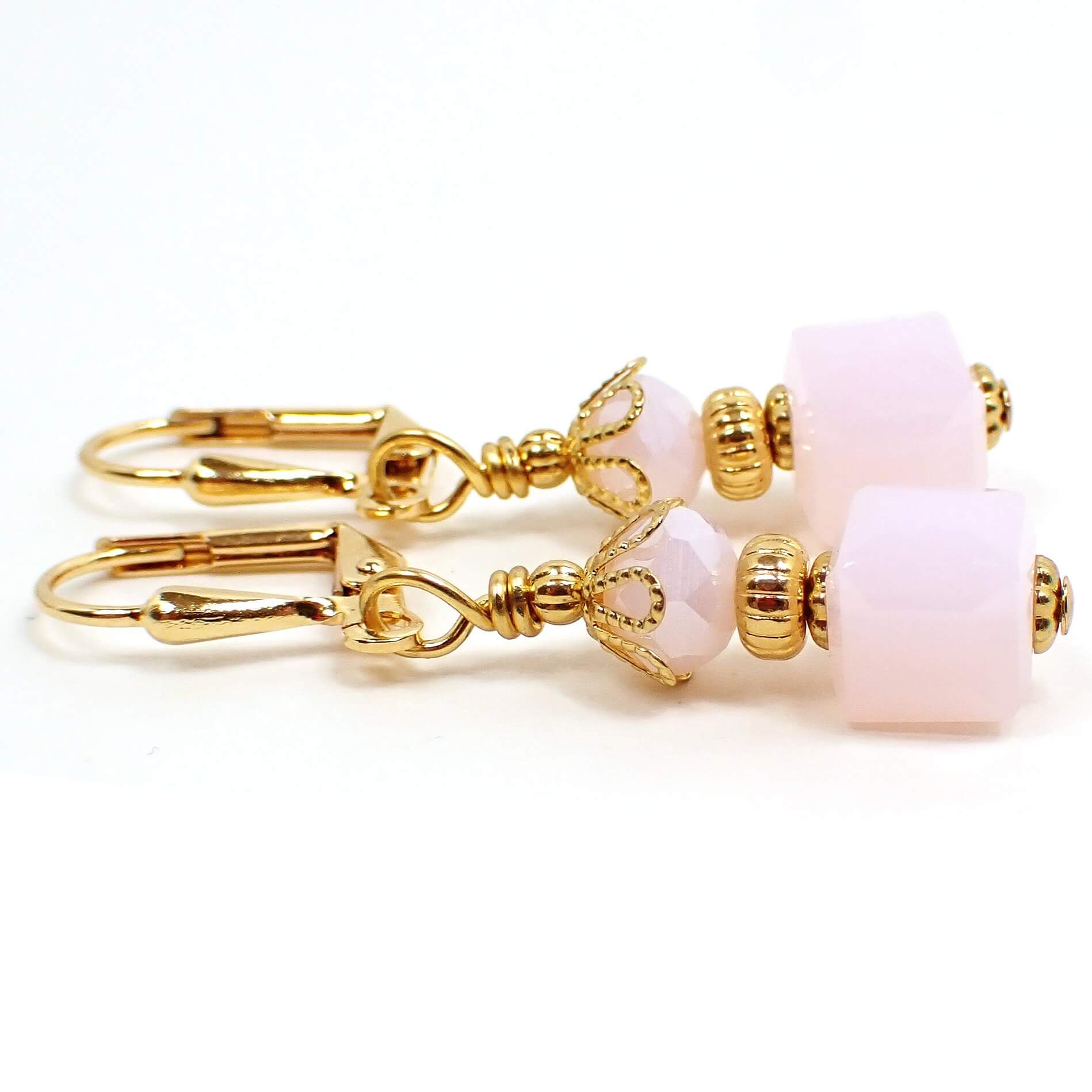 Side view of the small glass beaded earrings. The metal is gold plated in color. There are faceted glass crystal rondelle beads at the top and small cube shaped glass beads at the bottom. The beads are light pink in color.