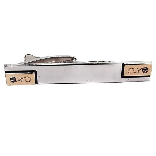 Front view of the Mid Century vintage Hickok tie clip. The majority of the metal is silver tone in color. There is a gold tone color rectangle at each end with an engraved floral design and a small clear rhinestone.