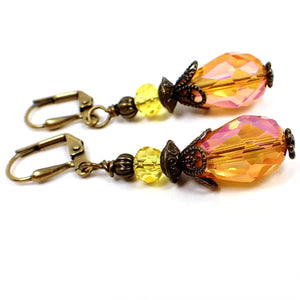 Angled side view of the handmade Pink Orange and Yellow vintage style teardrop earrings. The metal is dark antiqued brass in color. The top faceted bead is yellow and the bottom teardrop bead is orange and pink. Both beads are faceted glass crystal.