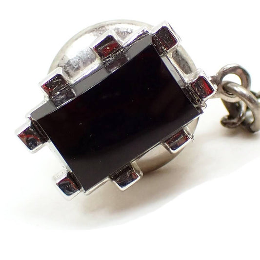 Enlarged front view of the Mid Century vintage Swank tie tack. The metal is silver tone in color and has a rectangle shaped design. There are two prongs coming out from each side. In the middle is a black glass cab.