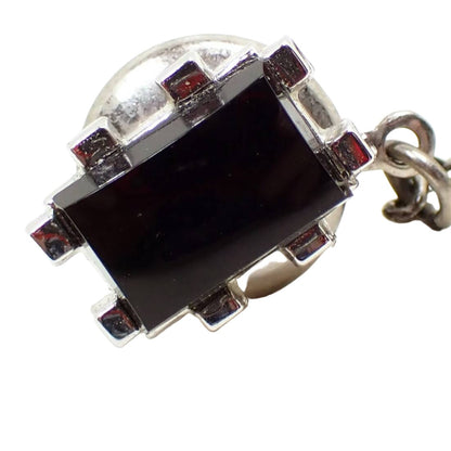 Enlarged front view of the Mid Century vintage Swank tie tack. The metal is silver tone in color and has a rectangle shaped design. There are two prongs coming out from each side. In the middle is a black glass cab.