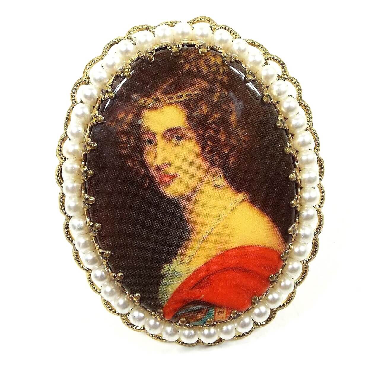Front view of the Mid Century vintage cameo brooch pin made in West Germany. It is oval in shape with a filigree edge and small round white plastic faux pearls. The metal is gold tone in color. There is a decal over a plastic cab on the front depicting a woman with curly brown hair with a white dress and red shawl. The background is dark brown. 