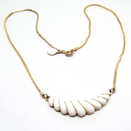 Front view of the retro vintage Avon pendant necklace. The metal is gold tone in color. The bottom pendant area is curved on top and scalloped on the bottom. There is a white plastic front with gold tone wire wrapping in between each scalloped area. 