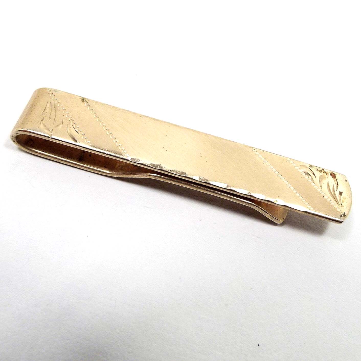 Angled front view of the Mid Century vintage etched tie bar. The metal is gold tone in color. There are diagonal areas of matte and shiny gold tone. At each end is an etched leaf like design. The bottom and top edge have a beveled texture.