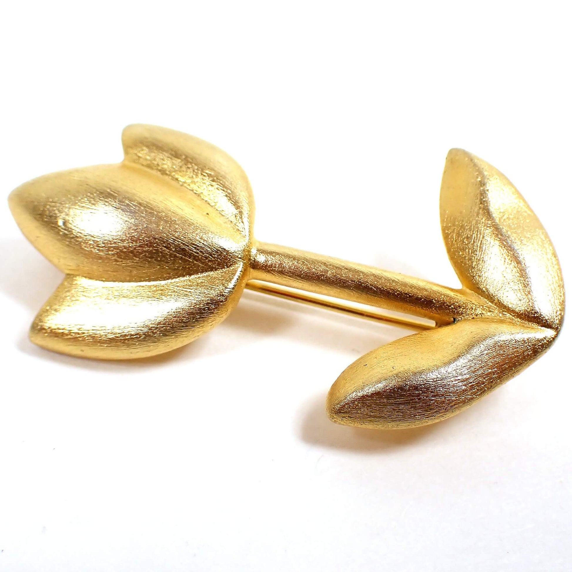 Angled front view of the retro vintage floral brooch pin. The metal is matte brushed gold tone plated in color. It is shaped like a flower with three petals going upward and two leaves at the bottom of the stem.