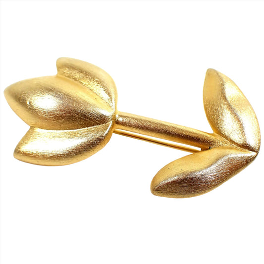 Angled front view of the retro vintage floral brooch pin. The metal is matte brushed gold tone plated in color. It is shaped like a flower with three petals going upward and two leaves at the bottom of the stem.