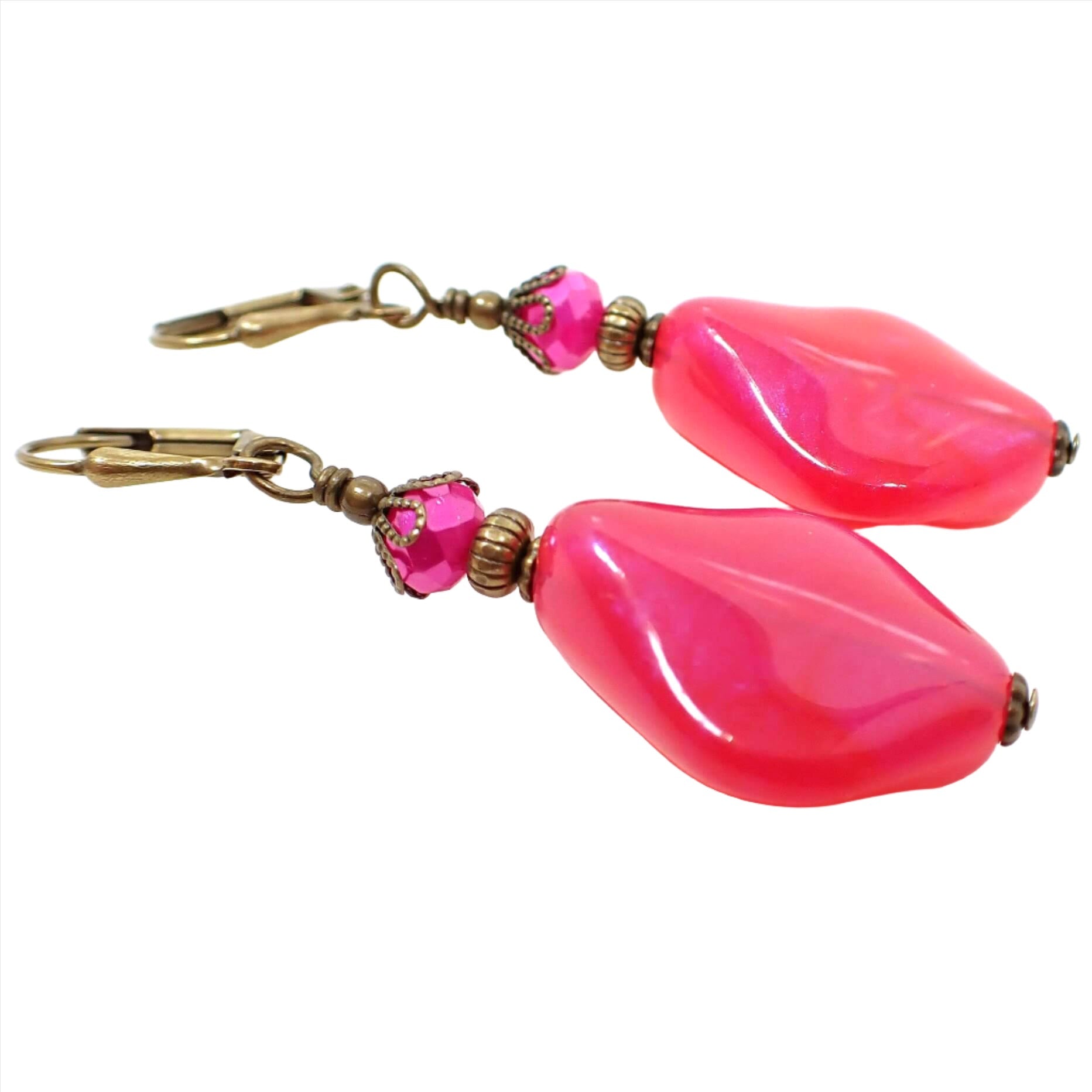 Angled side view of the handmade color shift earrings. The metal is antiqued brass in color. There are hot pink faceted glass crystals at the top. The bottom beads are shaped like curved and angled teardrops are are bright pink lucite with shimmers of purple color as you move around in the light.