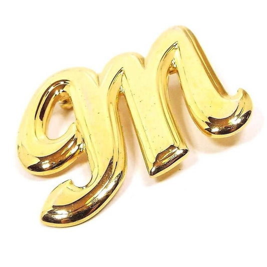 Angled front view of the retro vintage initial brooch pin. The metal is a bright yellow gold tone plated in color. It is shaped like a fancy block letter M.