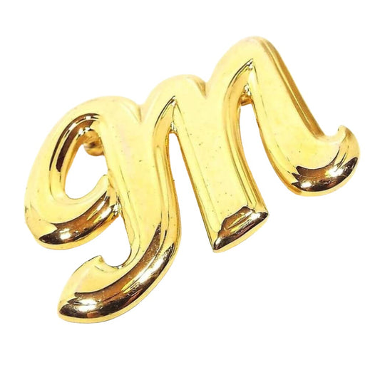 Angled front view of the retro vintage initial brooch pin. The metal is a bright yellow gold tone plated in color. It is shaped like a fancy block letter M.