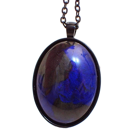 Enlarged front view of the large Goth handmade resin pendant necklace. The chain and pendant setting are black in color. The large oval pendant has a domed handmade resin cab with pearly black and deep blue resin.
